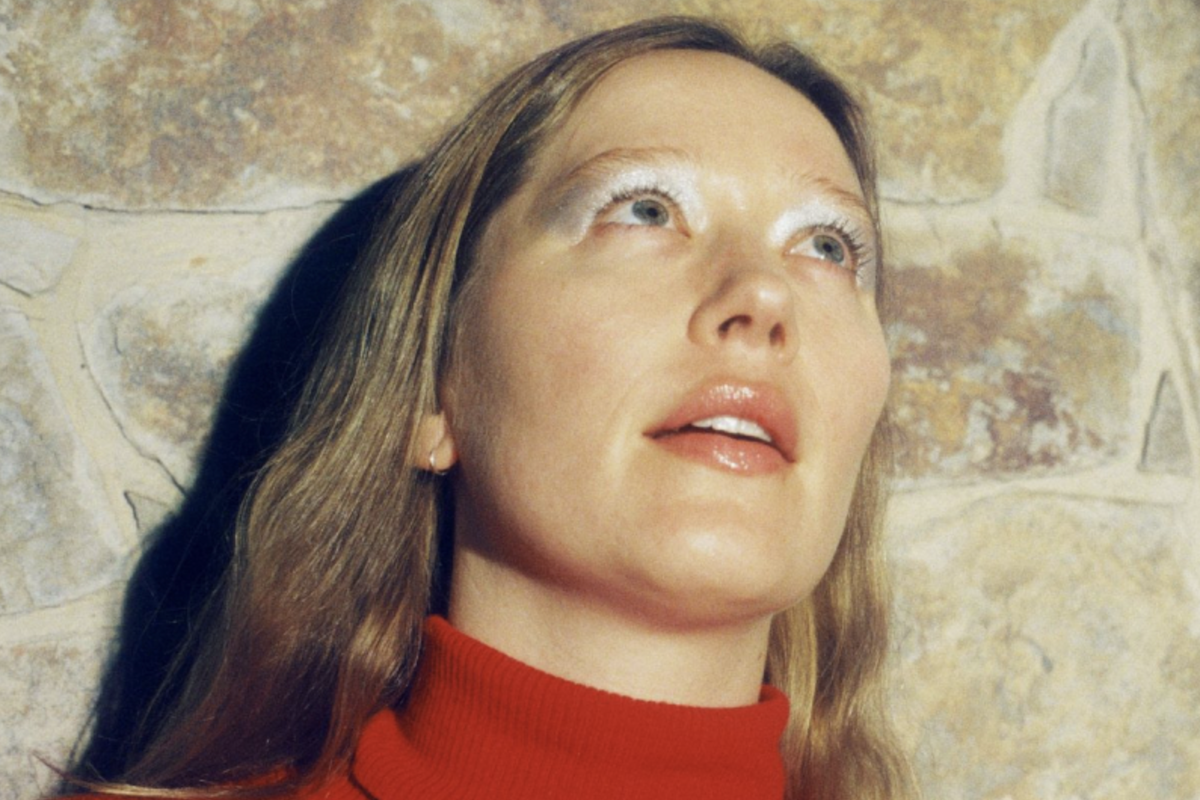 Close-up portrait of singer-songwriter Julia Jacklin in a red turtleneck sweater, wearing silver eyeshadow and looking upward against a stone wall.